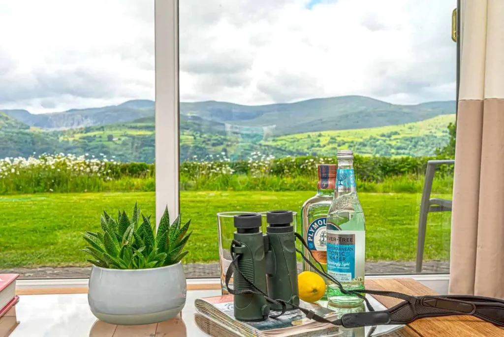 A close-up of a coffee table with binoculars and drinks, with the mountain view visible through the window, representing a quick transition from work to relaxation