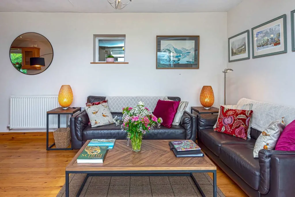The main lounge seating area at Laurel Cottage, showing a black leather sofa with colourful cushions and a wooden coffee table