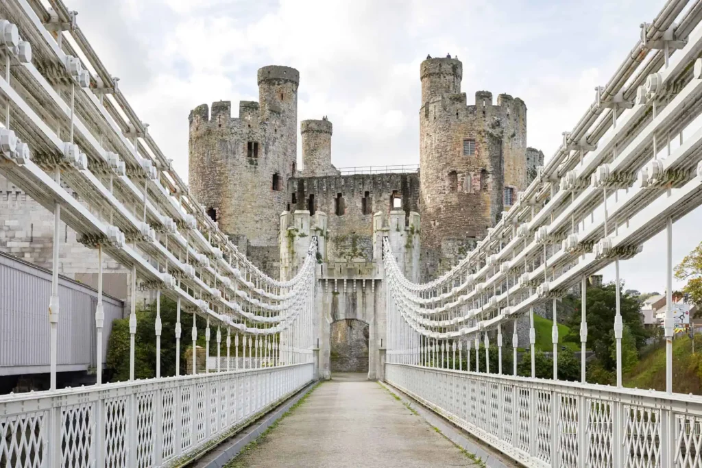 View of Conwy Castle from the entrance of the suspension bridge, with the bridge's walkway and iron support chains creating leading lines towards the fortress across the estuary.