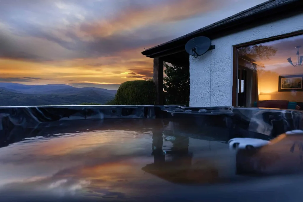 Conwy Valley Getaway | home hero hot tub sunset Hot tub at water level at sunset, Conwy Valley and mountains reflected in the water, cottage glowing in background